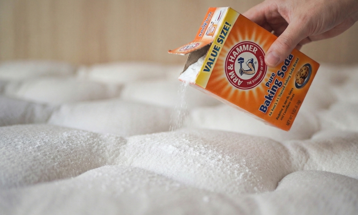 A bare mattress with an even dusting of baking soda across the surface, a small sieve and bowl nearby, soft morning light in a simple bedroom, realistic photo