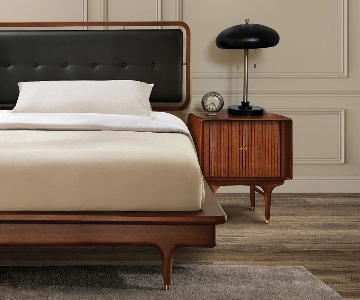 A bedroom nightstand vignette with a brass table lamp, a medium wood nightstand, and a lighter wood bed frame in the background, soft evening light, real photography