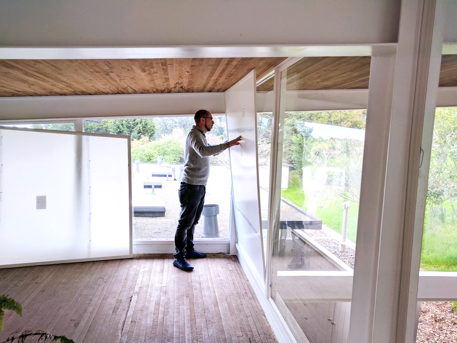 A bedroom window in daylight with a clear removable acrylic window insert fitted inside the frame, with a neatly made bed nearby, real home photo