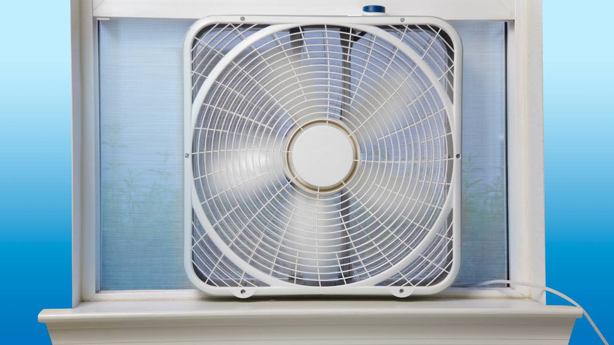 A bedroom window open with a box fan positioned to blow air outward, sunlight hitting a neatly made bed and upholstered headboard
