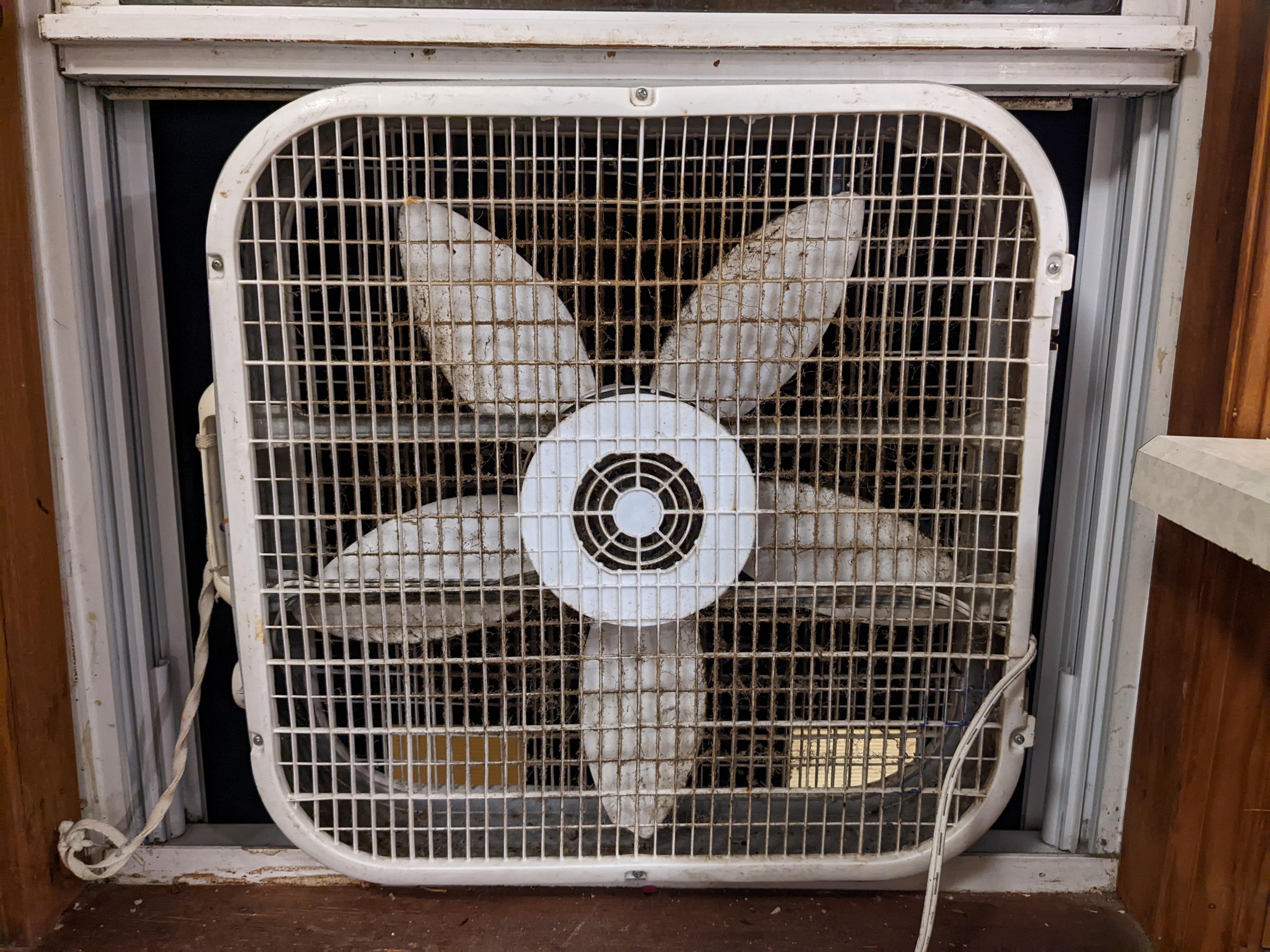 A box fan placed in an open apartment window facing outward to exhaust indoor air, with sunlight coming into an empty room