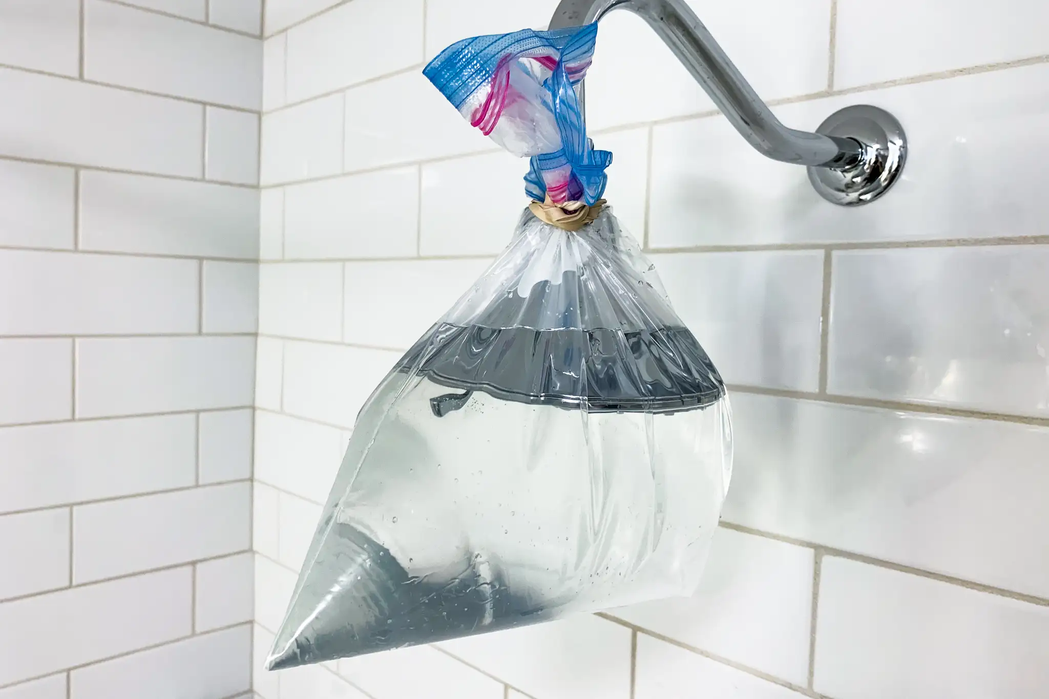 A chrome showerhead wrapped in a clear plastic bag filled with vinegar and tied at the neck, hanging inside a clean tiled shower, realistic photo