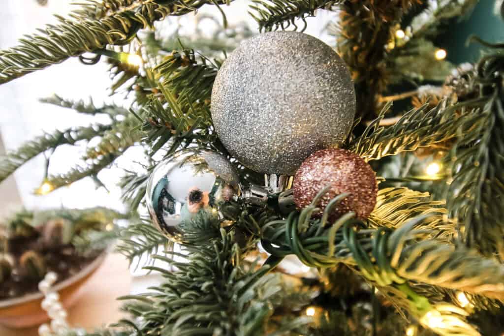 A close-up of a Christmas tree with ornaments arranged in a small cluster of three in complementary colors, warm white lights glowing in the background