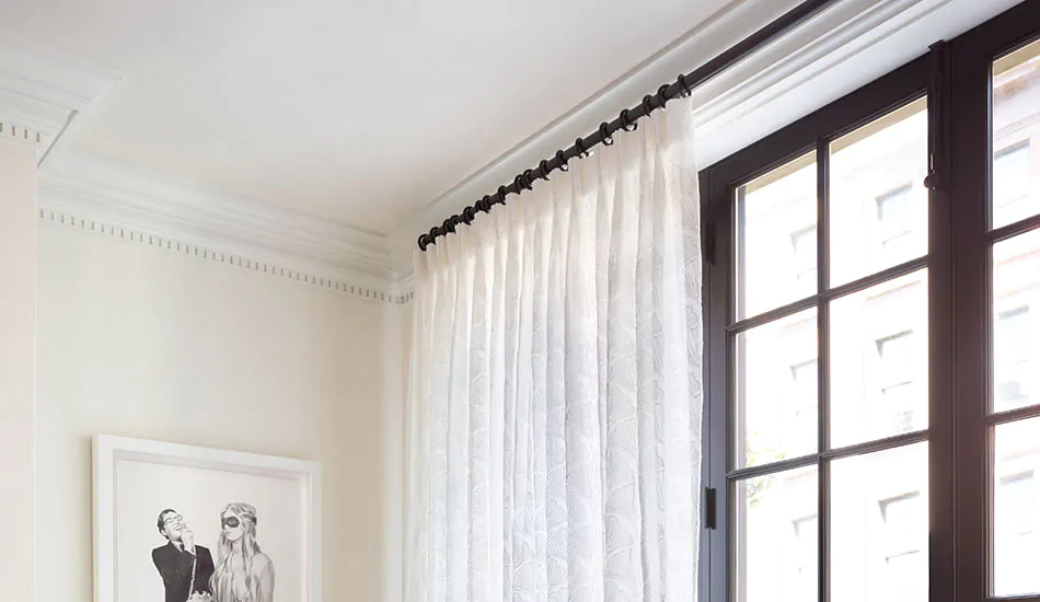 A close-up of a curtain rod mounted just below white crown molding above a window, with brass brackets and light fabric panels hanging straight