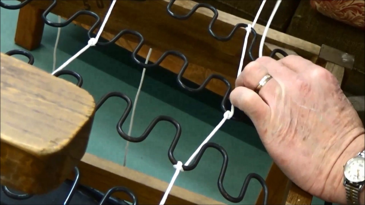 A close-up of a sofa underside showing zigzag springs and a person tightening a spring clip screw with a screwdriver