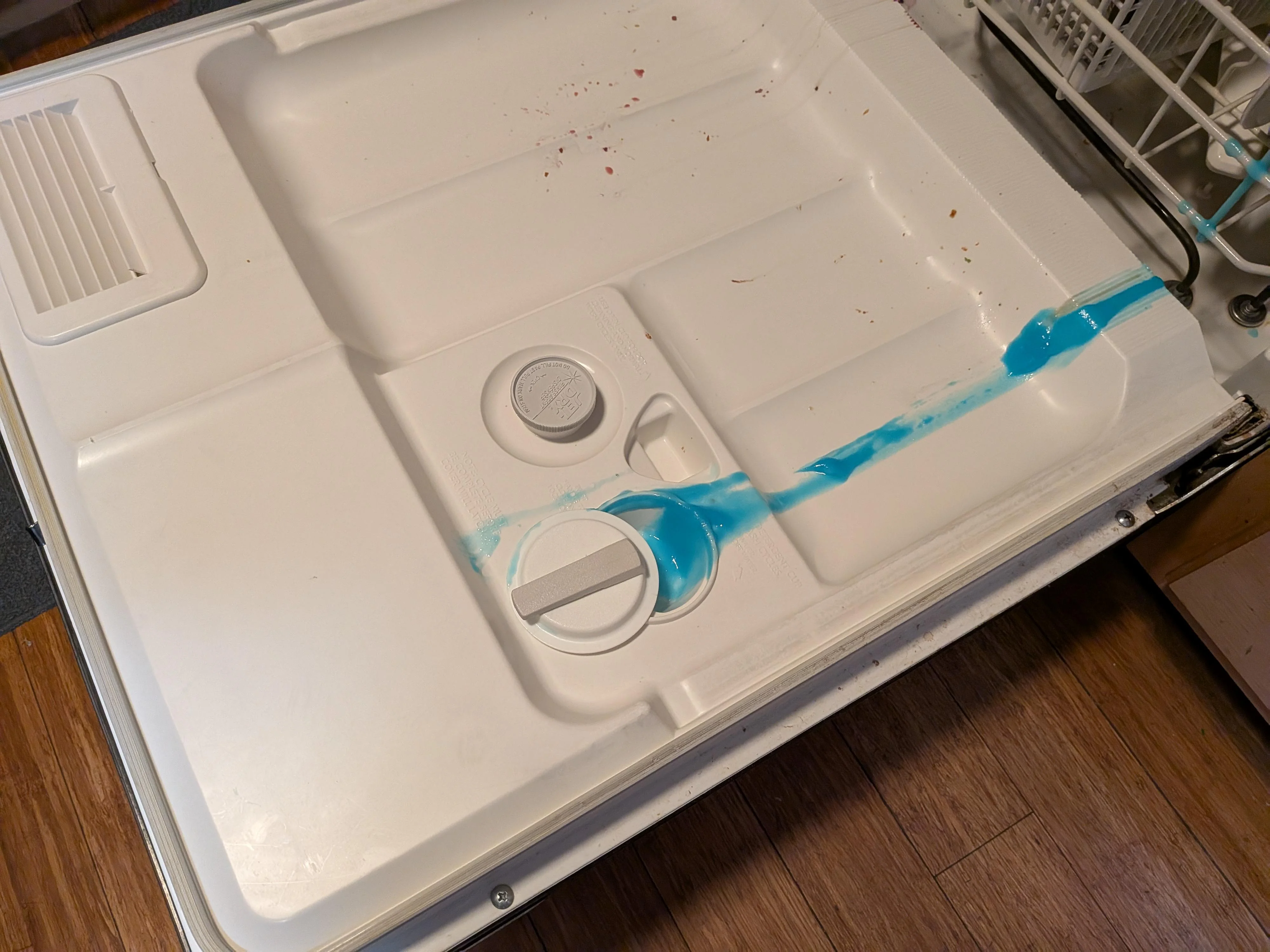A close-up photo of a dishwasher detergent dispenser being scrubbed with a small brush, showing mild residue around the cup