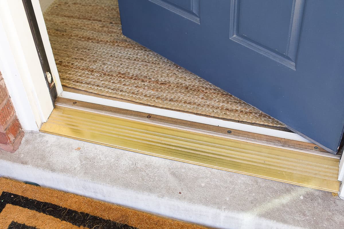 A close-up photo of a front door threshold and weatherstripping where the door meets the sill, showing the rubber seal and metal threshold in natural daylight
