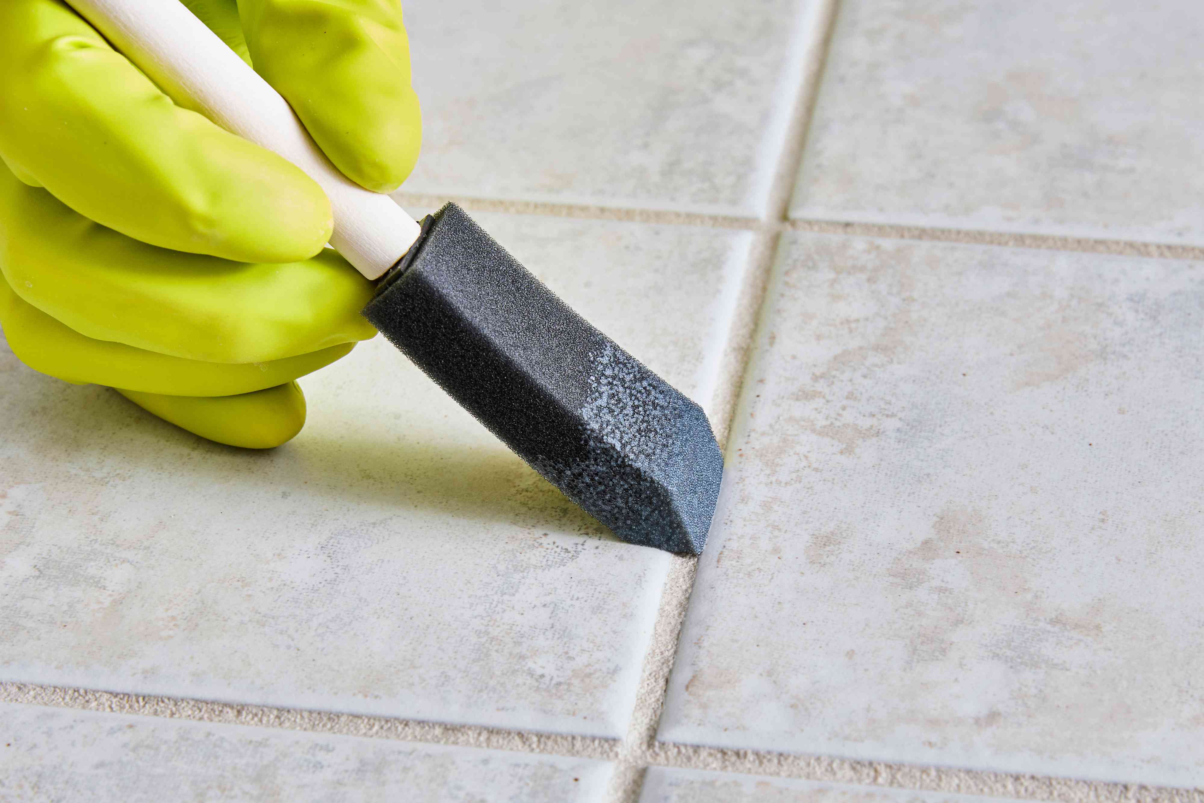 A close-up photo of a hand using a small foam brush to apply clear penetrating grout sealer along grout lines on a bathroom floor, soft natural light