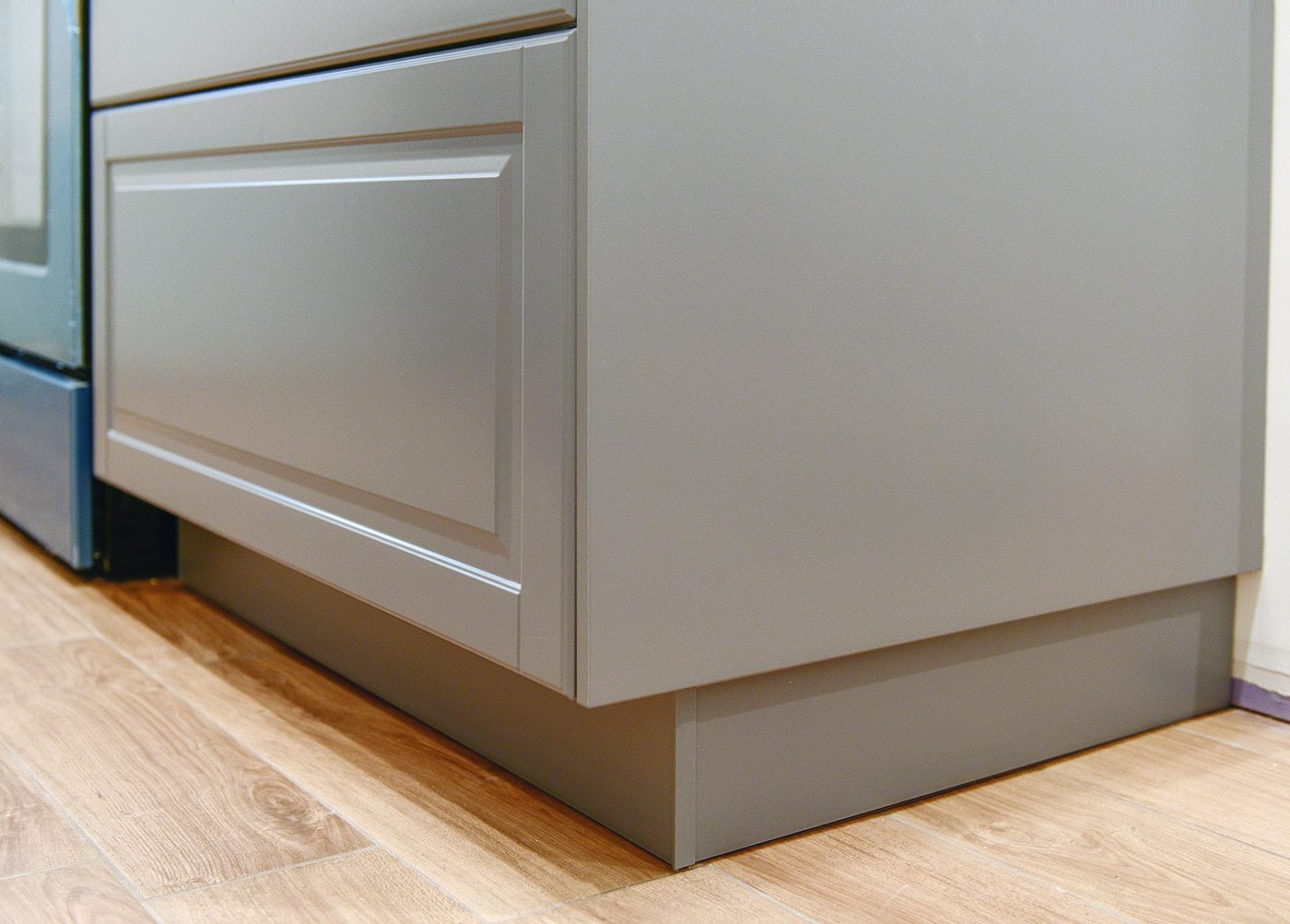A close-up photo of a kitchen toe-kick and base trim detail in a neutral color, showing a clean junction between cabinet base and floor