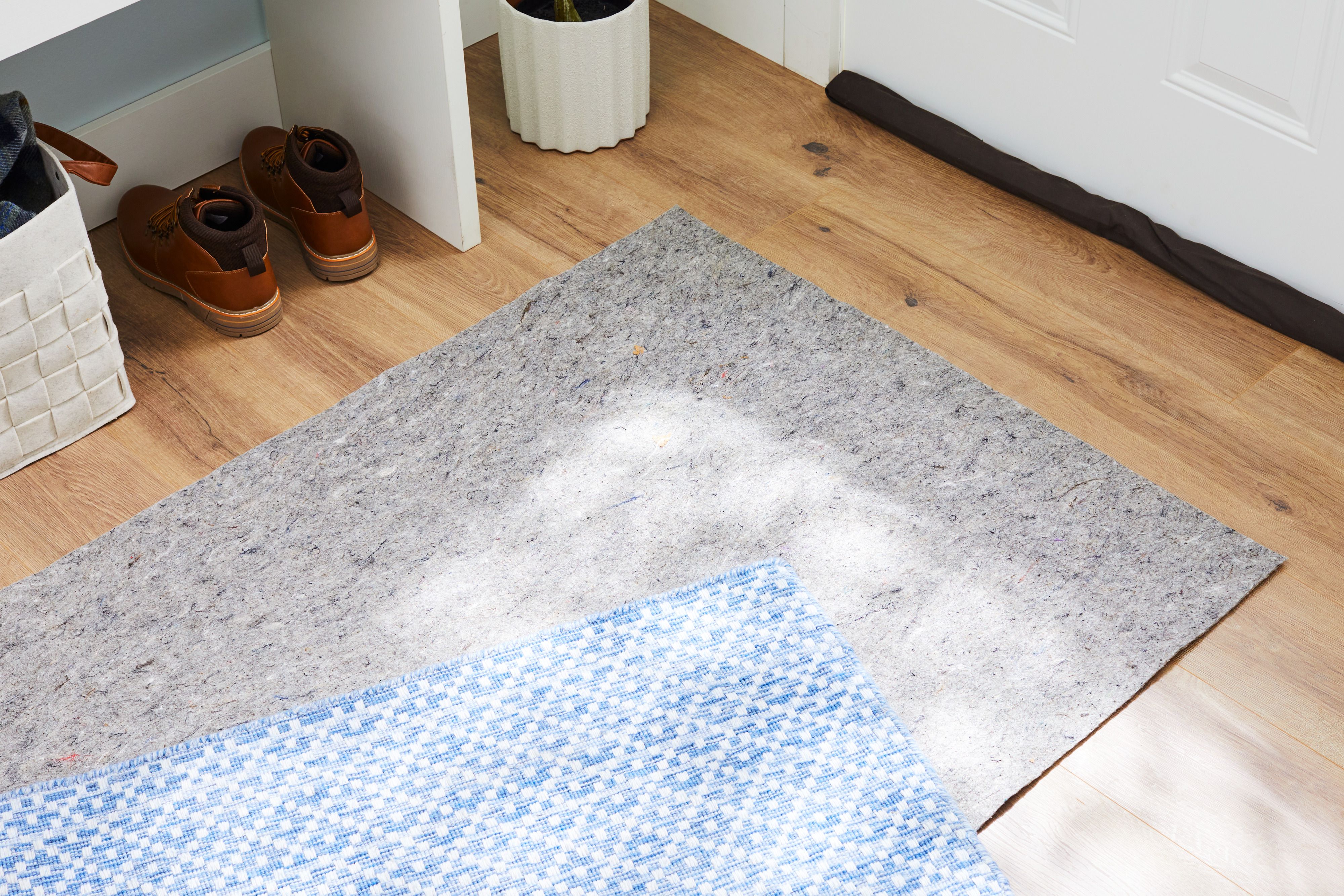 A close-up photo of a patterned area rug corner lifted to show a felt and rubber rug pad on a warm-toned hardwood floor