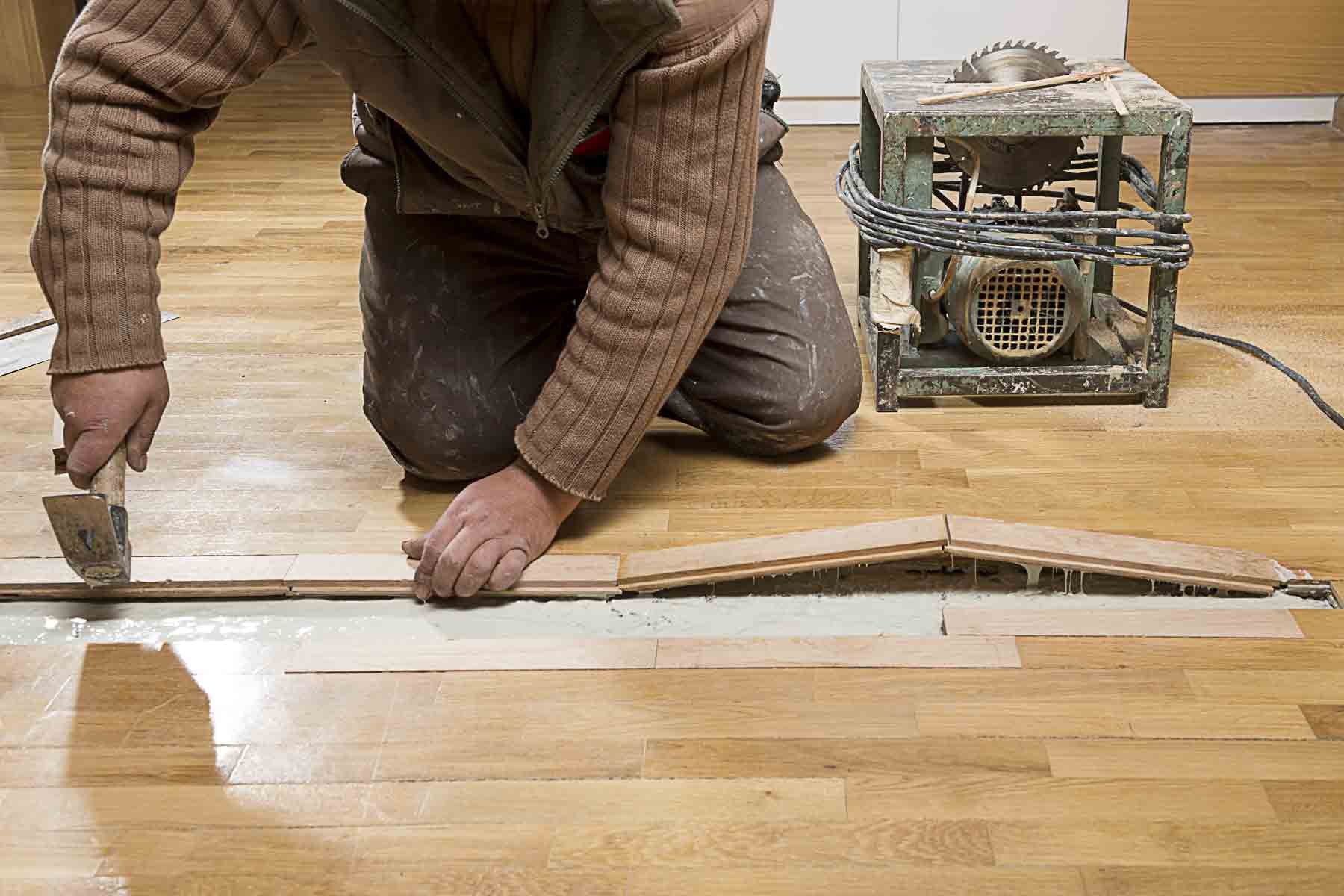 A close-up photo of a person using a drill driver to sink a small trim-head screw into a hardwood floor near a wall, with painter’s tape marking the suspected joist line