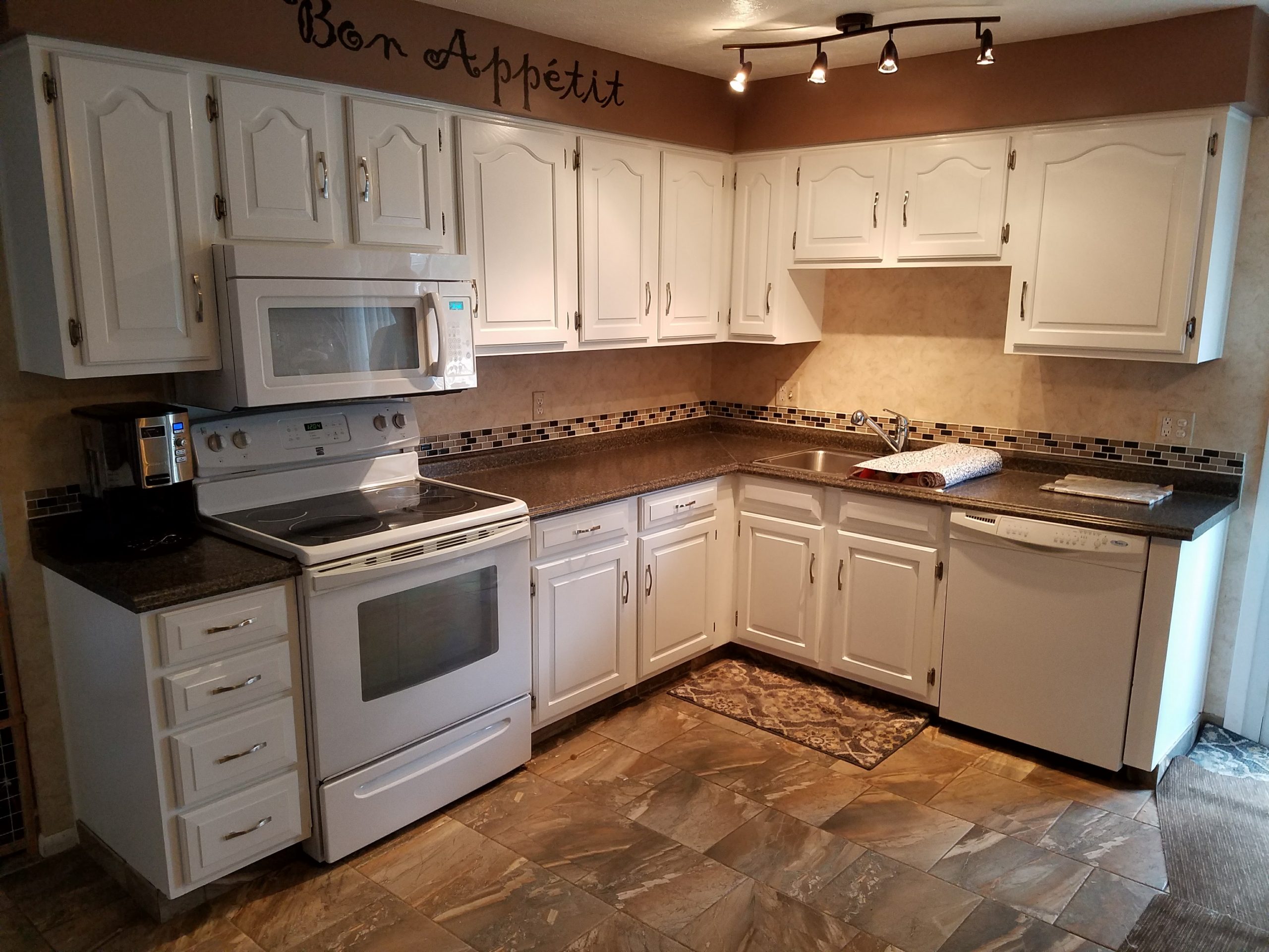 A close-up photo of a small paintbrush applying touch-up paint to a cabinet edge near a hinge, with painter’s tape protecting the surrounding area