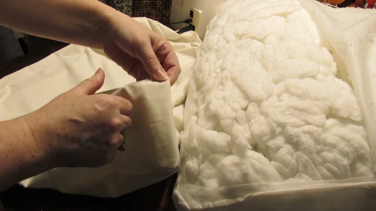 A close-up photo of hands adding loose polyester fiberfill into a back cushion insert on a sofa, with the insert opening visible and sunlight coming through a nearby window