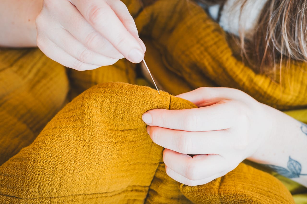 A close-up photograph of a blunt needle being inserted beside a small snag in beige woven upholstery, with the fabric held steady by fingertips
