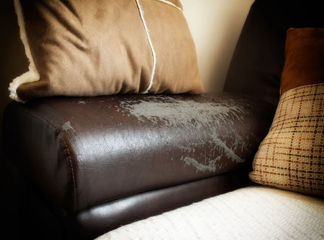 A close-up photograph of a dark brown bonded leather sofa seat cushion with visible peeling, flaking, and cracked surface texture in natural window light