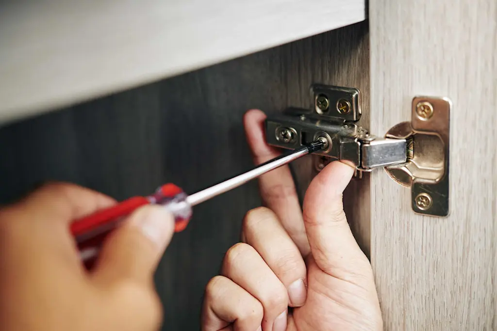 A close-up photograph of a hand using a screwdriver to adjust a concealed European cabinet hinge inside a light-colored kitchen cabinet