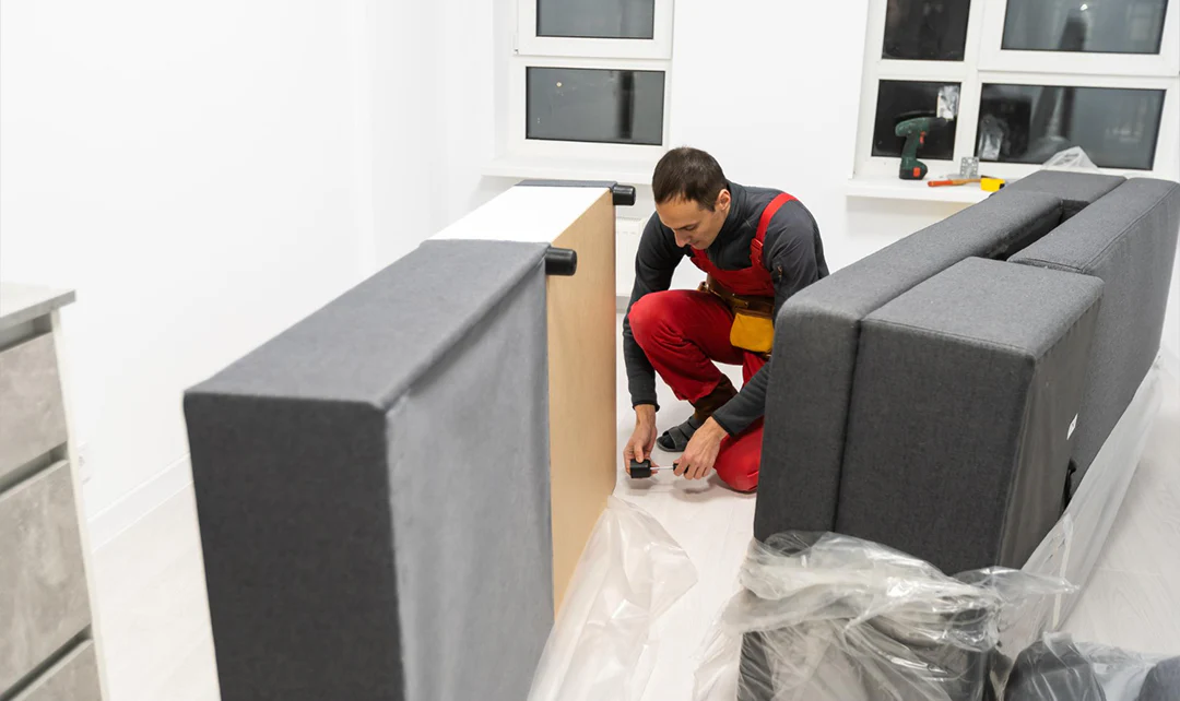 A close-up photograph of a manual recliner flipped forward on a soft rug, showing the metal reclining mechanism and linkage arms while a person inspects it with a flashlight in a cozy living room