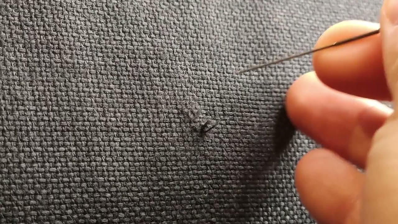 A close-up photograph of a neutral woven upholstery fabric on a sofa arm with a small cat snag and a hand holding a needle nearby in soft window light