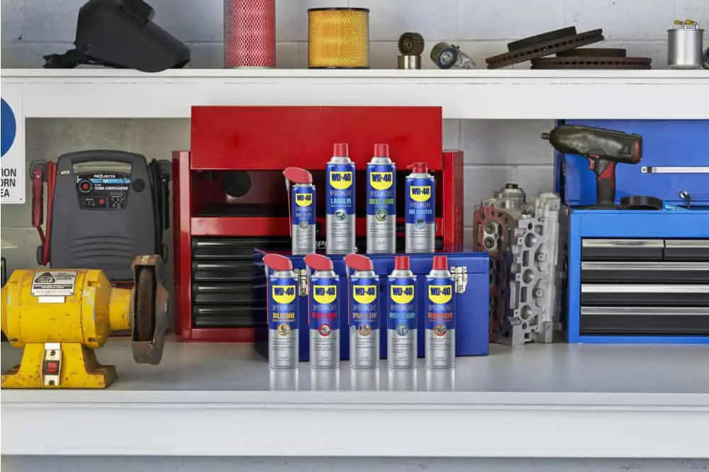 A close-up photograph of a person applying a small amount of white lithium grease to a recliner metal pivot hinge using a narrow straw nozzle, with the chair tipped forward