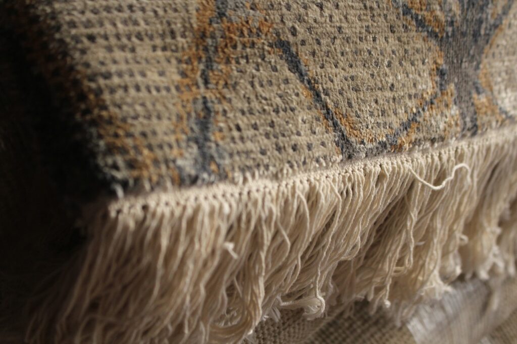 A close-up photograph of a wool area rug edge with cream fringe, showing a few tassels fraying and loose warp threads near the binding on a hardwood floor, natural window light