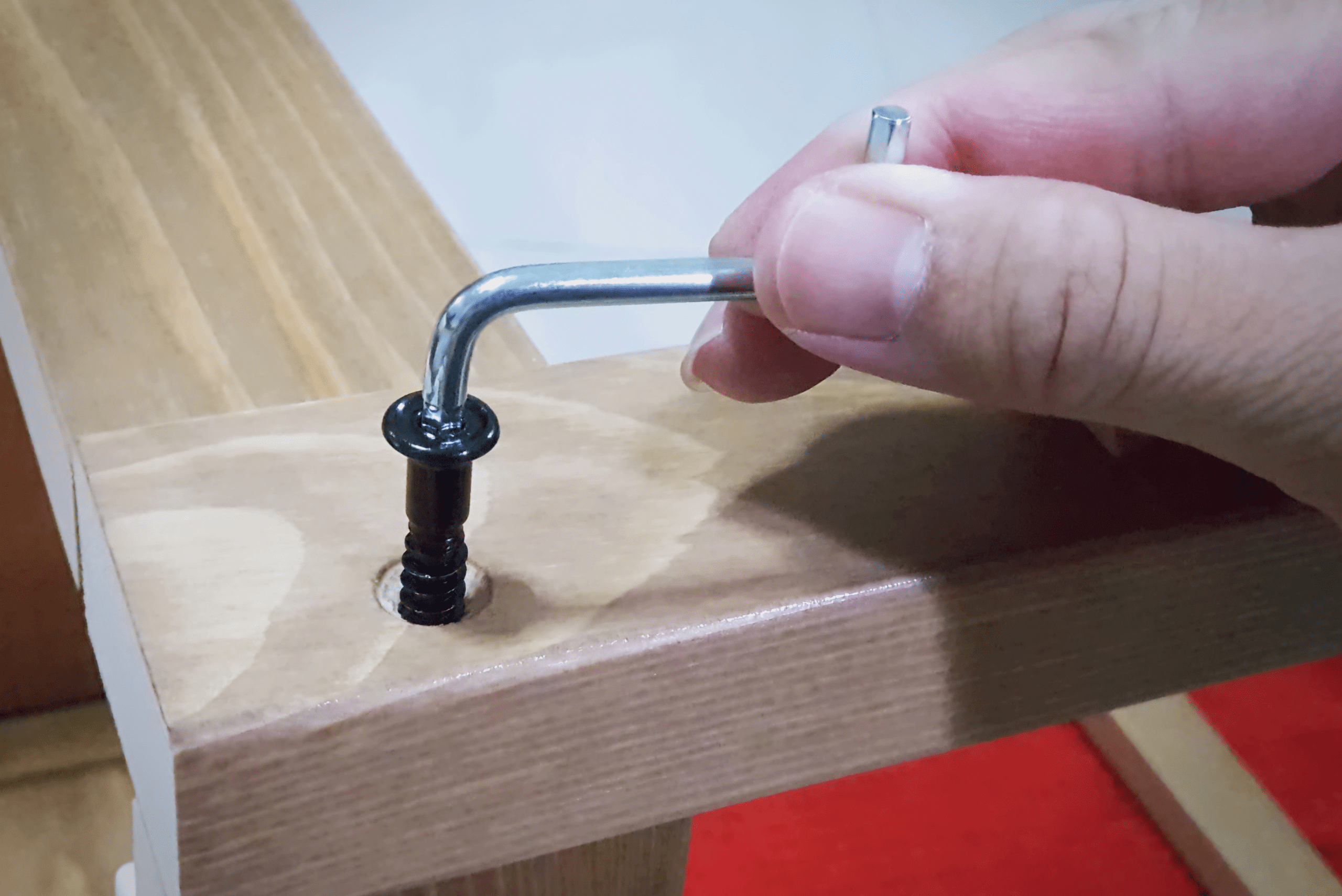A close-up real photograph of hands tightening a dining chair seat bolt with a hex key while the chair is upside down on a soft blanket