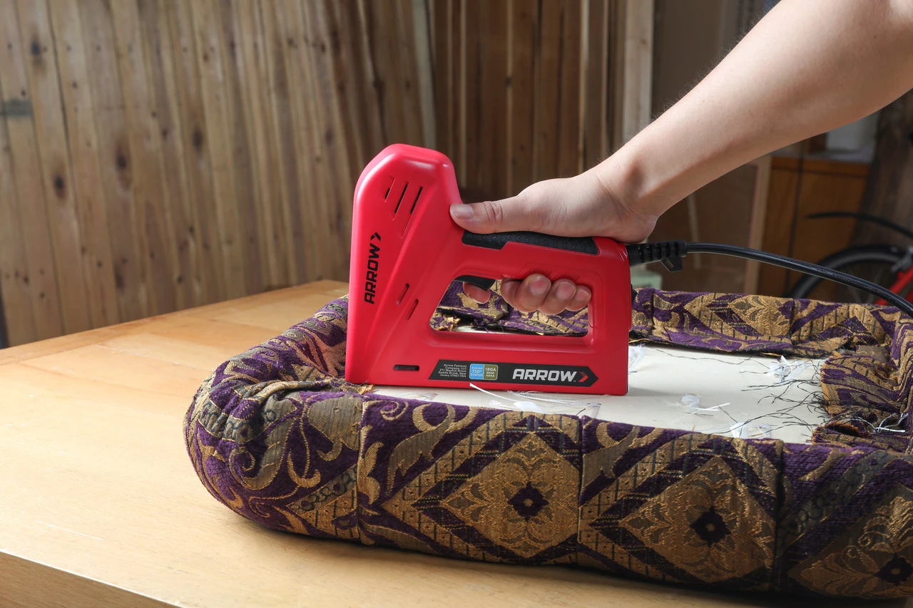 A close view of a hand holding an upholstery stapler near the back edge of a headboard frame, with fresh staples and fabric folded neatly