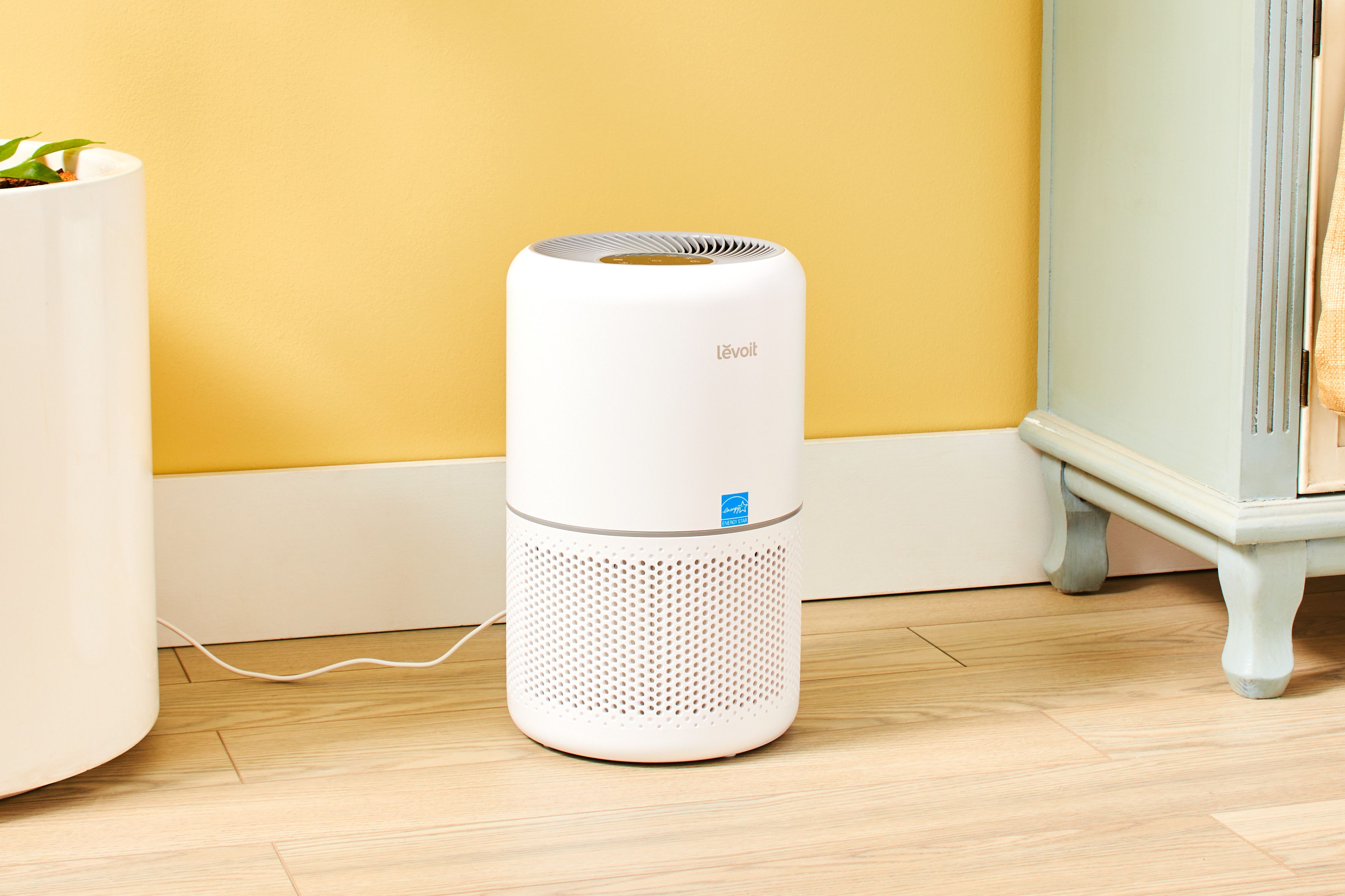 A cylindrical air purifier running on a hardwood floor next to a newly delivered fabric sofa in a calm neutral living room, realistic home photography