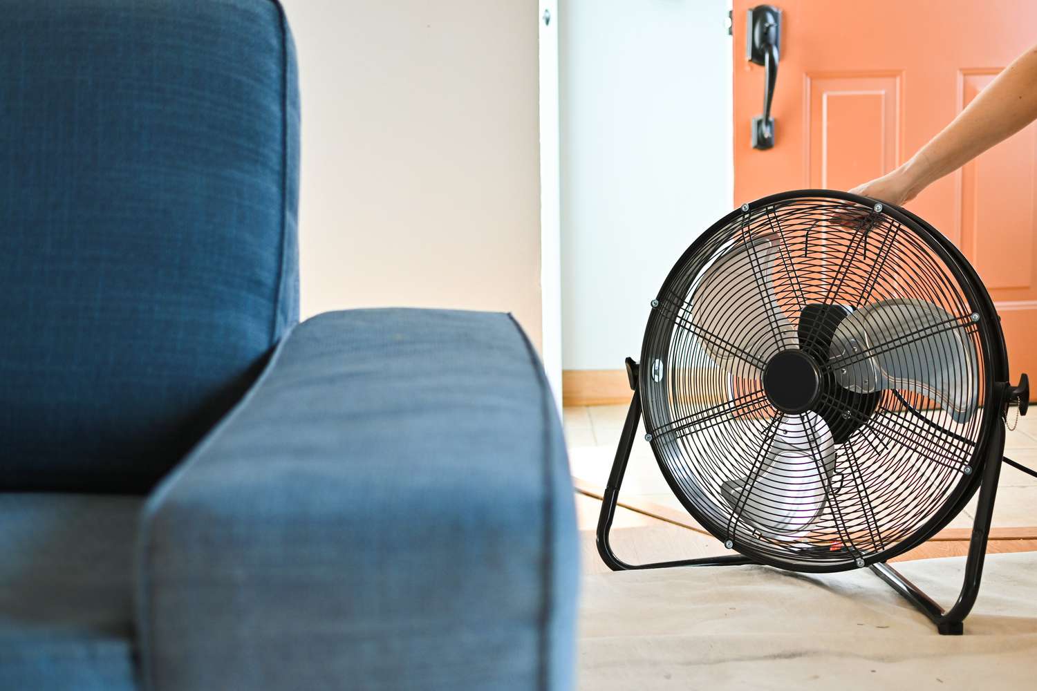 A fabric sofa drying in a sunlit room with an oscillating fan placed a few feet away and an open window letting in fresh air