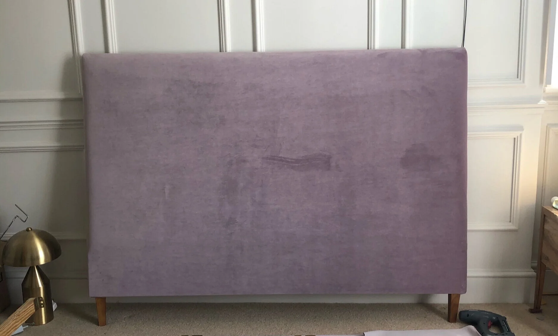 A fabric-upholstered headboard laid face-down on a clean floor with the back dust cover partially removed, exposing staples and a thin layer of old foam