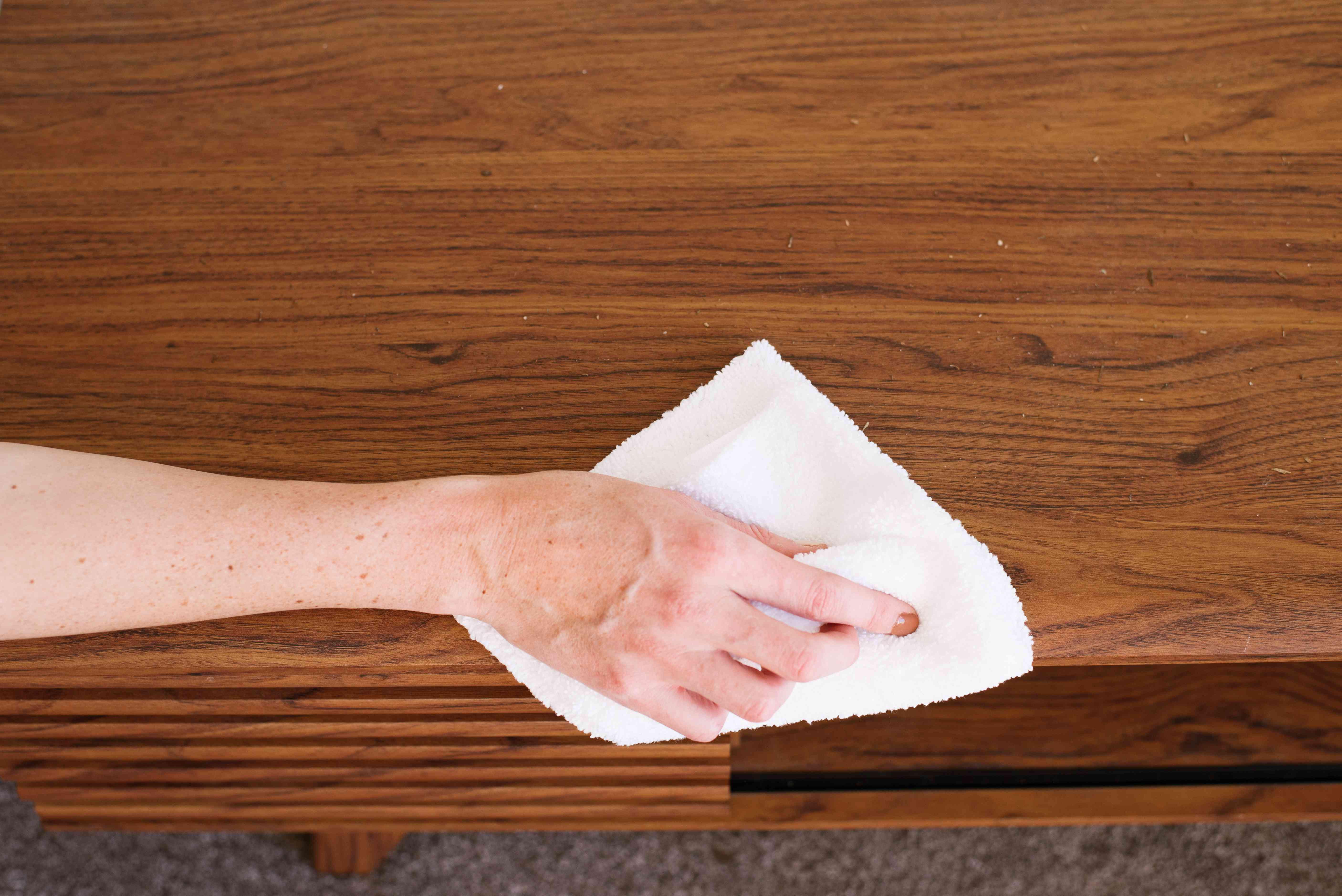 A hand buffing a small hazy patch on a finished wooden dining table using a clean microfiber cloth in warm indoor lighting
