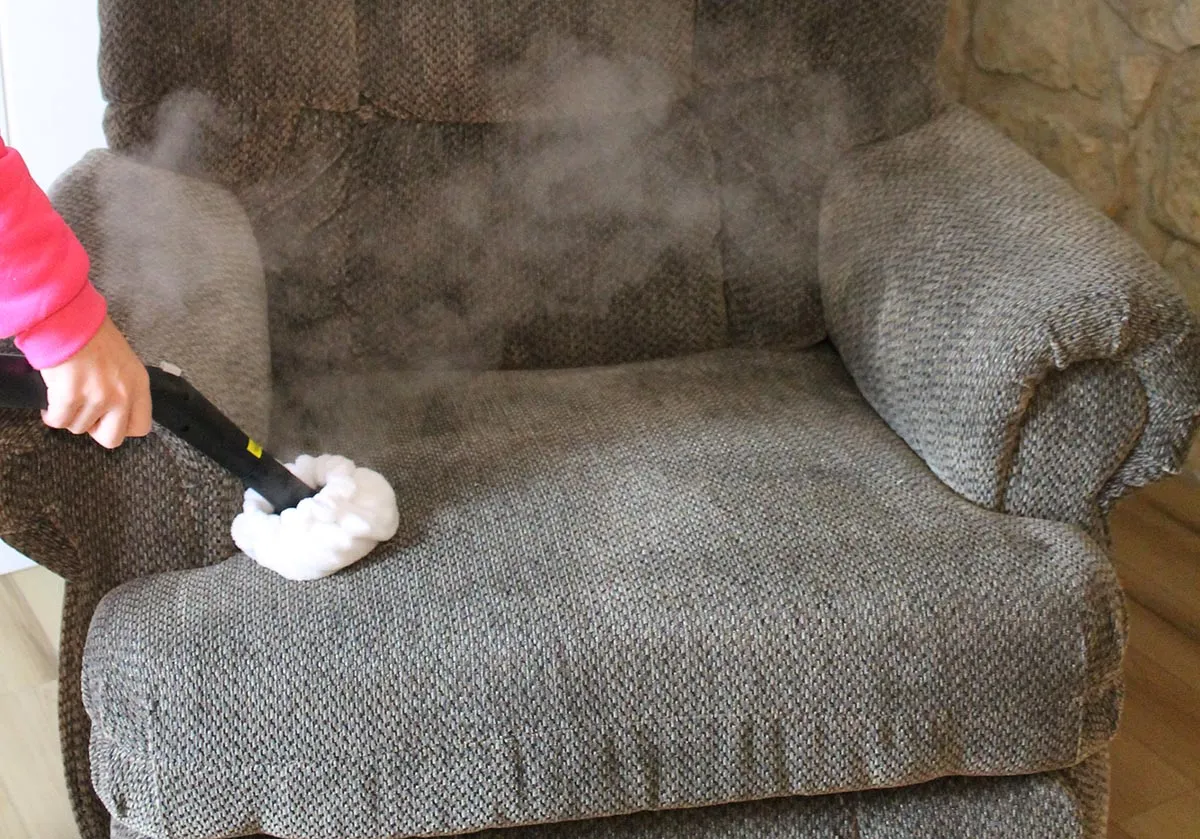 A handheld garment steamer being used carefully on the arm of a light gray fabric sofa, with gentle steam visible in natural window light