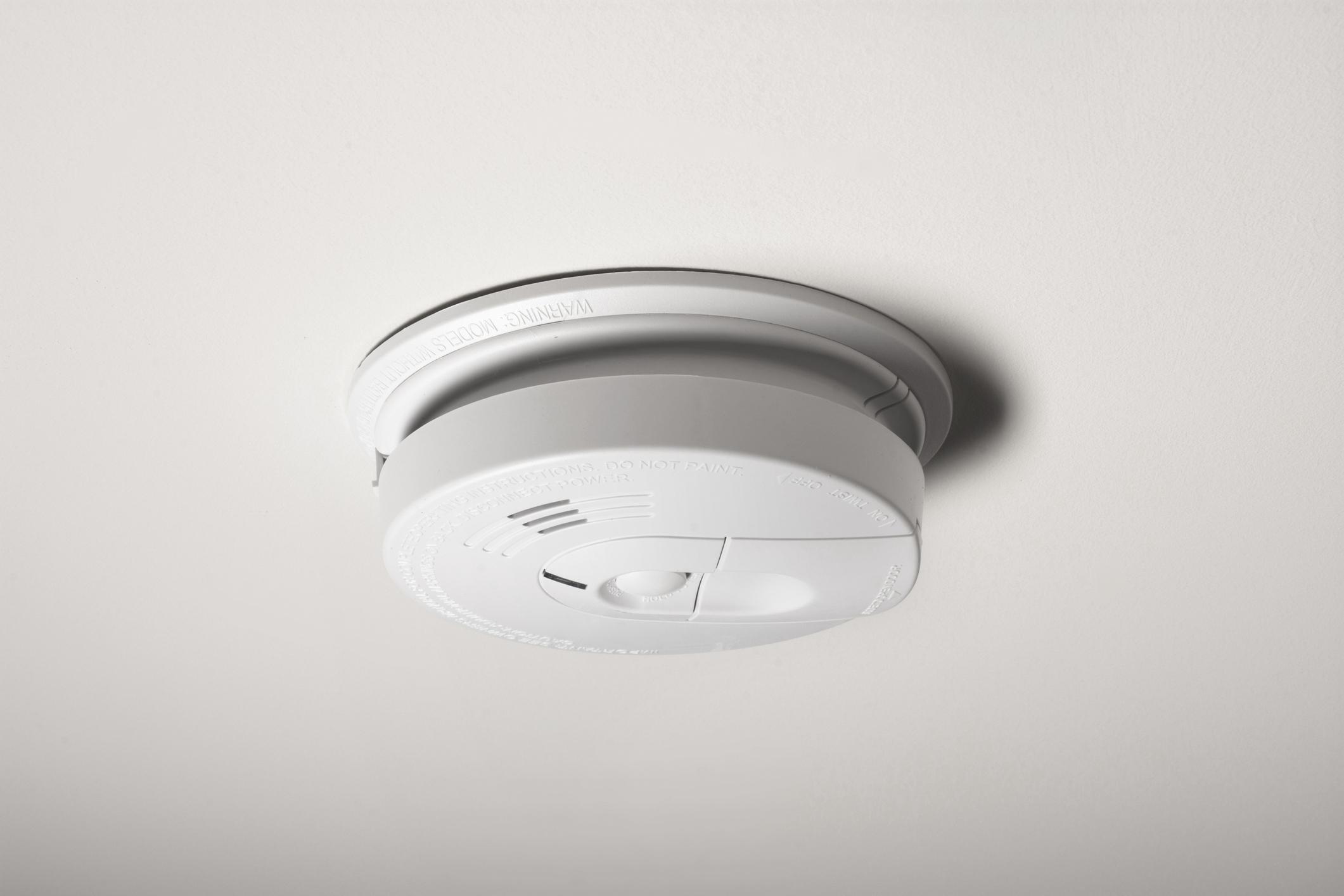 A hardwired smoke alarm mounted on a white ceiling with a small gap showing the base plate, realistic home maintenance photo