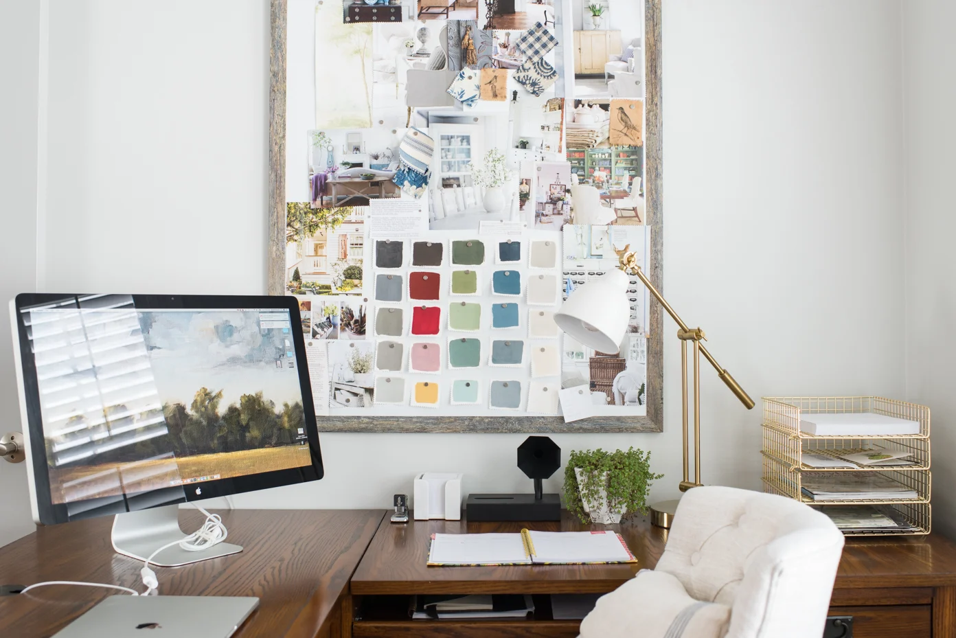 A home office with a large linen pinboard on the wall filled with photos and swatches, a simple desk below, and organized supplies on a narrow shelf