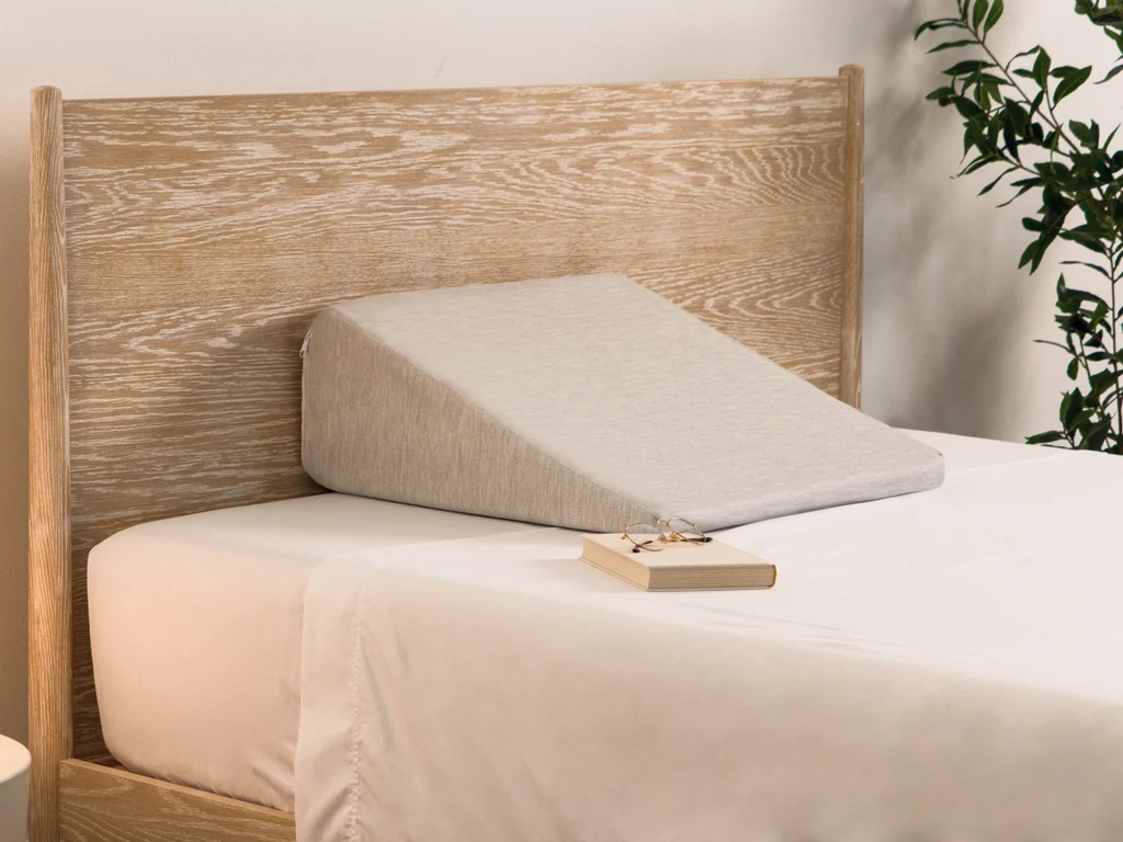 A long neutral foam wedge tucked between a mattress and a beige upholstered headboard, mostly hidden behind stacked sleeping pillows