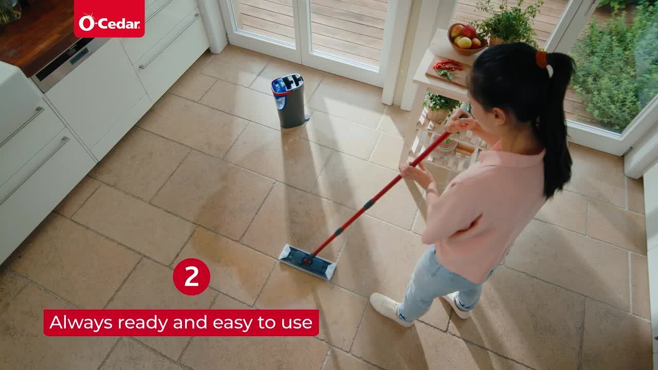 A microfiber flat mop being wrung out over a bucket on a porcelain tile hallway floor in a small apartment, with natural window light