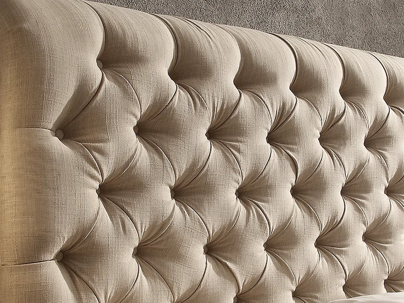 A neutral fabric upholstered headboard pulled slightly away from a bedroom wall to show the narrow gap behind it, with daylight coming in from a nearby window