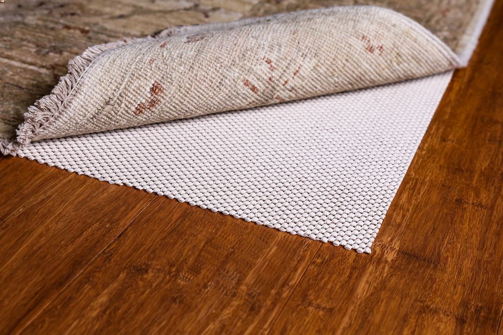 A new area rug partially lifted to show a thick rug pad underneath on a wood floor, photographed in natural window light