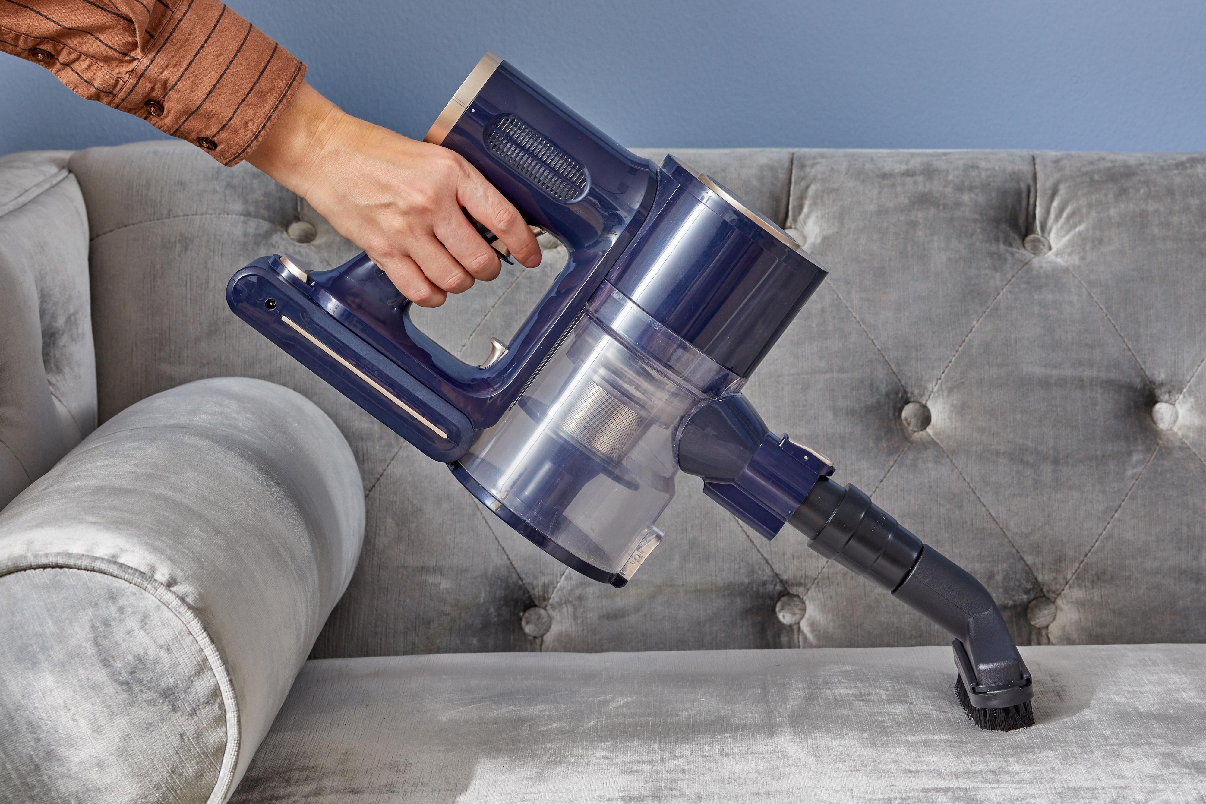 A person gently vacuuming a deep green velvet sofa with a soft upholstery brush attachment in a sunlit living room