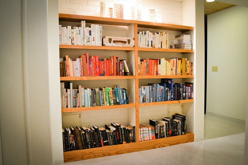 A person holding a tape measure across a blank wall in a home, pencil marks on painter's tape indicating planned bookshelf widths and heights, natural daylight