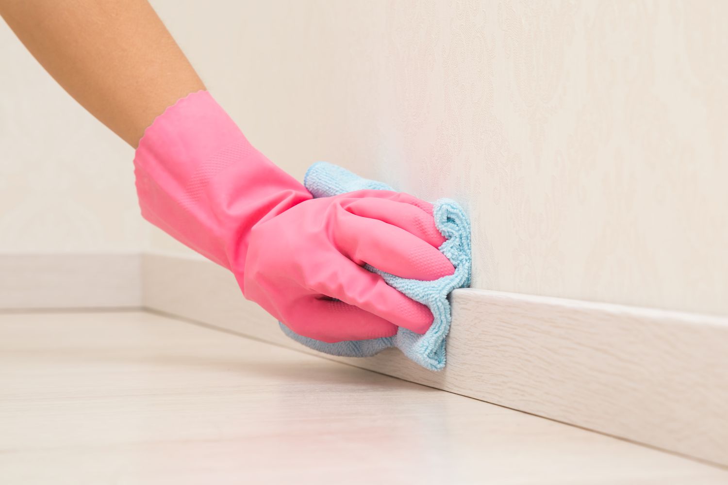 A person kneeling on a hardwood floor wiping white baseboards with a damp microfiber cloth while a small dog sits nearby, natural window light, real home photo