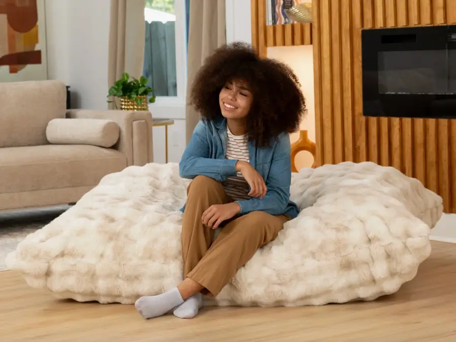 A person kneeling on a rug while fluffing an oversized floor cushion with both hands in a warm, softly lit living room