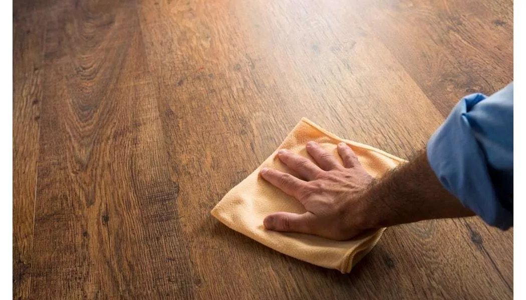 A person kneeling to buff luxury vinyl plank flooring with a dry white microfiber cloth in a sunlit living room