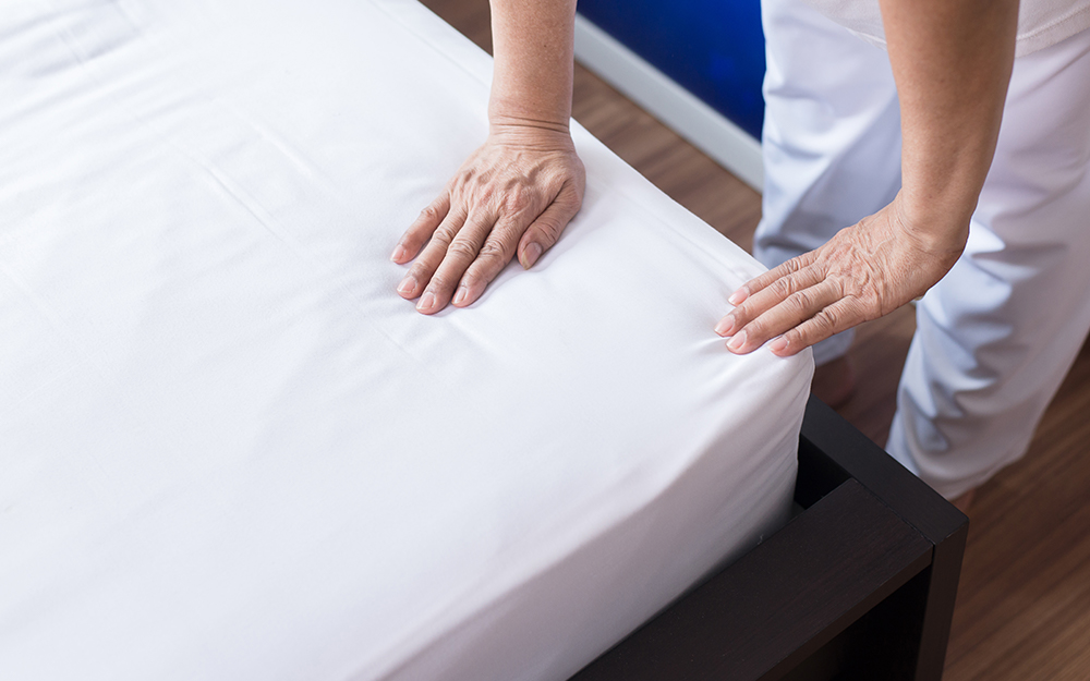 A person smoothing a fitted sheet over a neatly made bed in a calm bedroom, showing the mattress layers underneath subtly through the fabric
