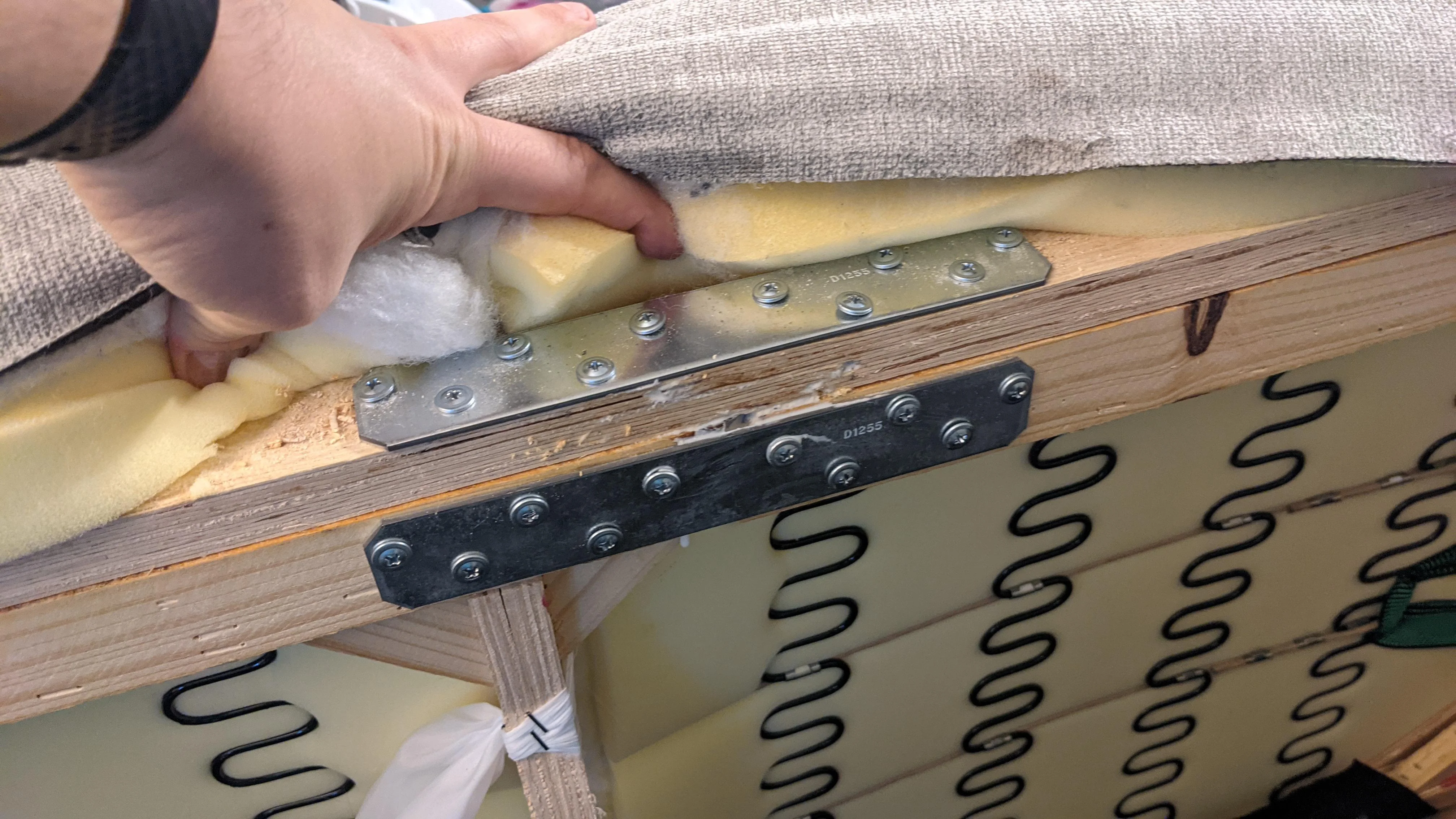 A person tipping a sofa forward in a living room to access the underside, using a socket wrench to tighten a loose frame bolt, natural window light