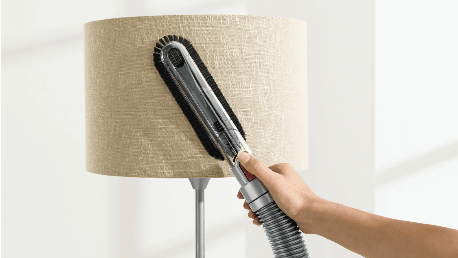 A person using a vacuum upholstery brush on low suction to gently clean a white fabric lampshade on a wooden table, realistic indoor photo