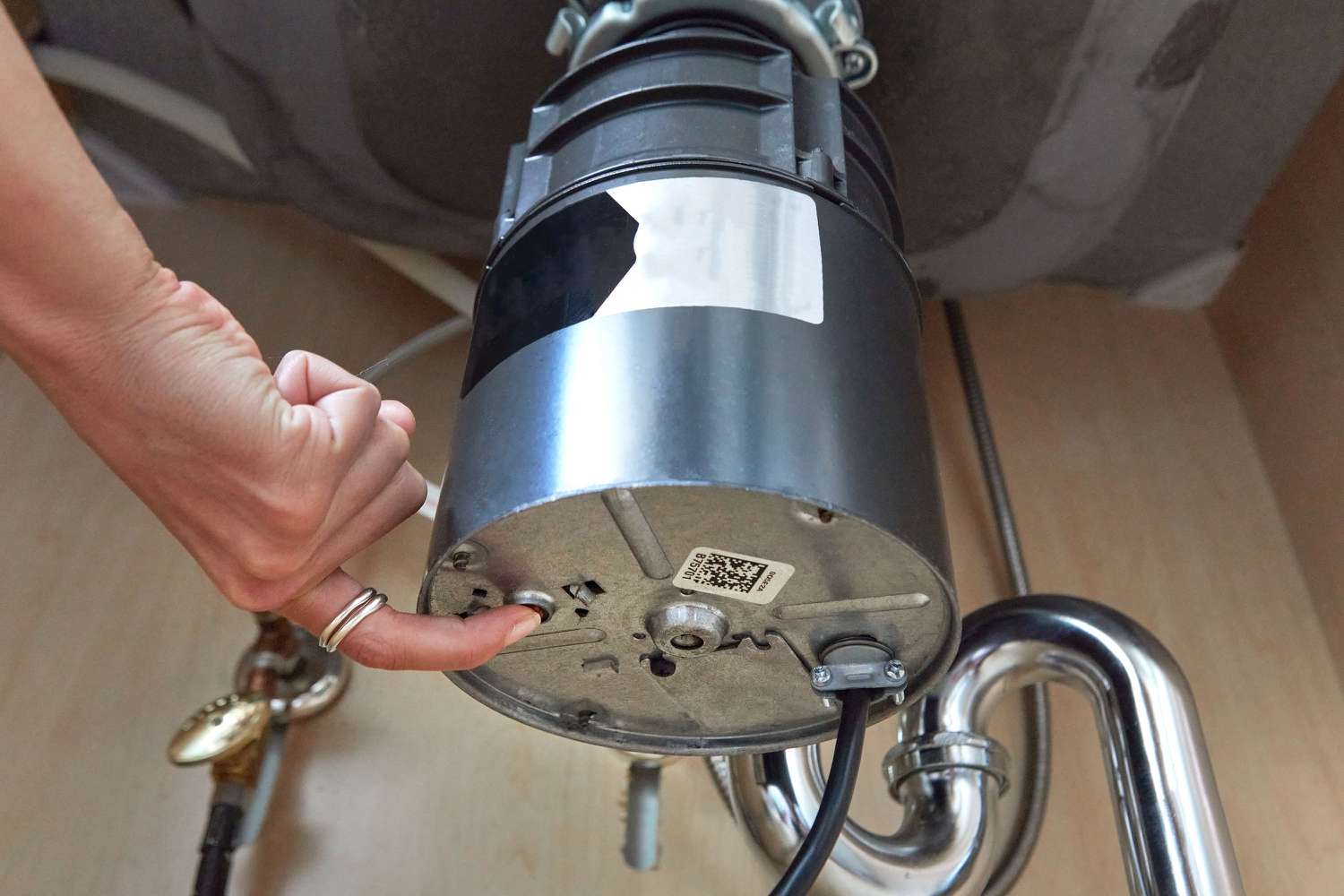 A person’s hand pressing the reset button on the underside of a garbage disposal under a kitchen sink, realistic photo