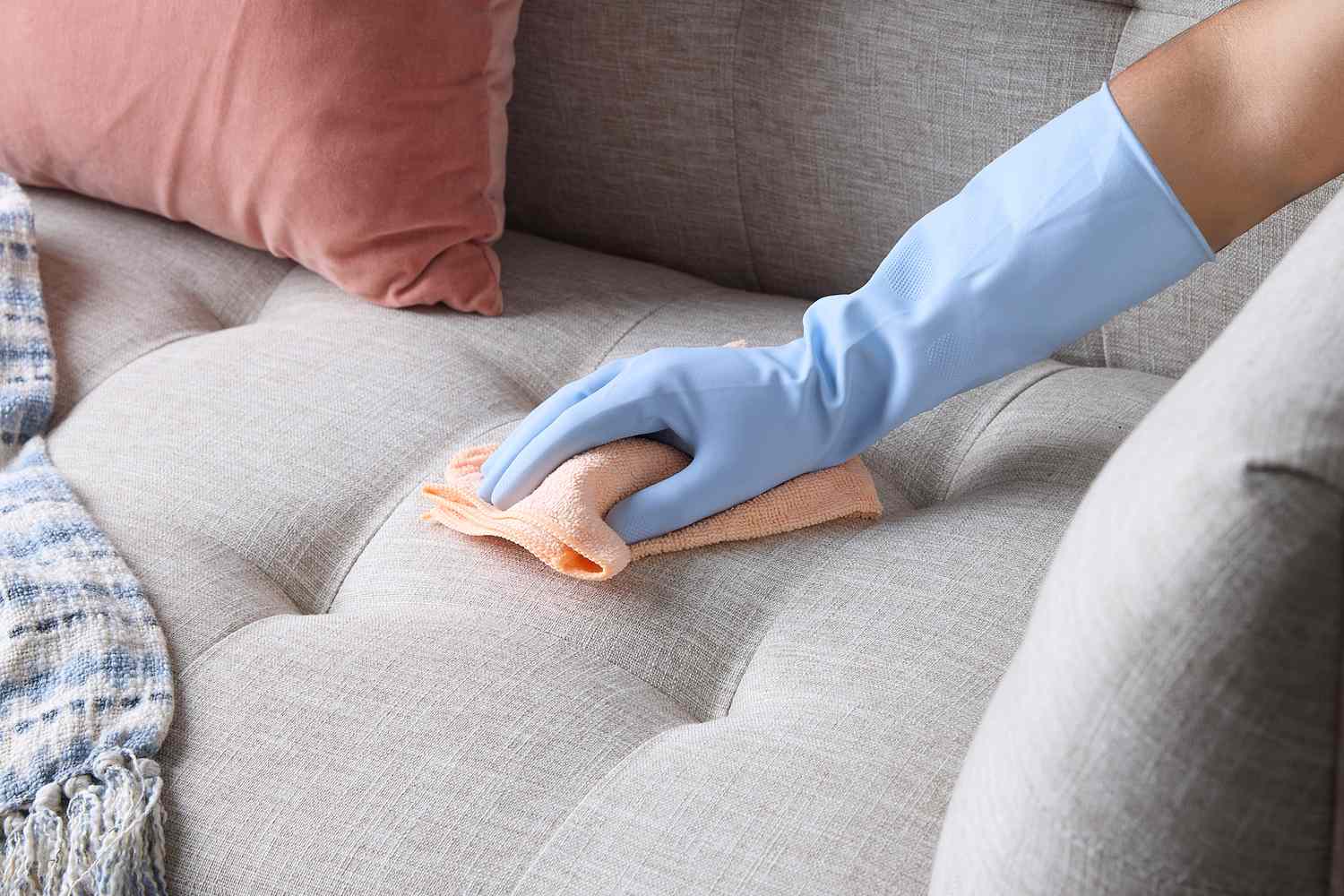 A photo of a hand using a cotton swab to apply a small amount of hydrogen peroxide to a tiny stain on a light cream upholstered sofa, close-up with soft daylight