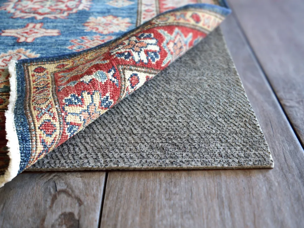 A photo of an area rug corner lifted to show a dark rug pad underneath that looks slightly damp on a hardwood floor