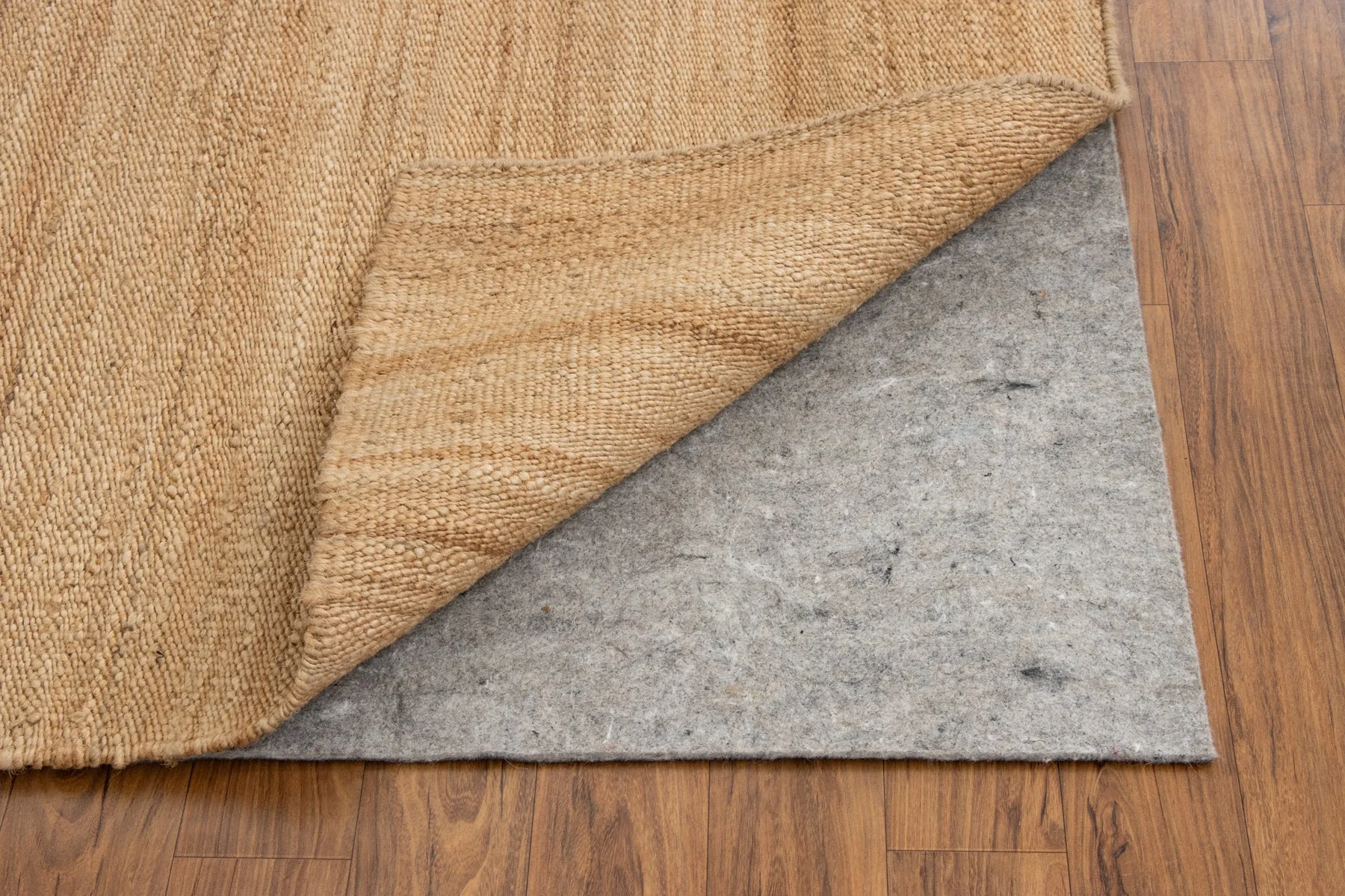 A photo of the corner of an area rug lifted to show a rug pad underneath on a hardwood floor, warm indoor lighting