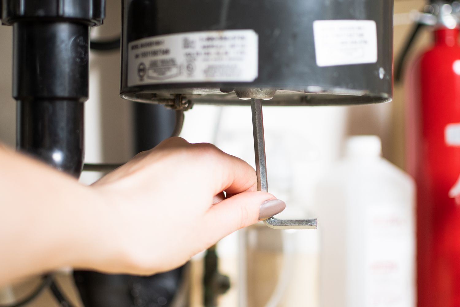 A photo under a kitchen sink showing an Allen wrench inserted into the bottom hex socket of a garbage disposal, with a hand turning it